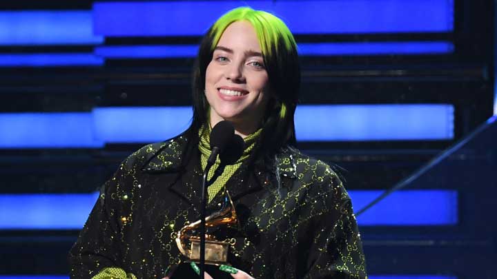 Billie Eilish, wearing a black and green dress matching her hair, holding an award on stage with a microphone, symbolizing her journey with Tourette Syndrome.