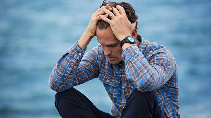 Understand the complexities of A distressed man sitting on the floor with crossed legs, holding his head, wearing a checkered shirt, symbolizing Generalized Anxiety Disorder.