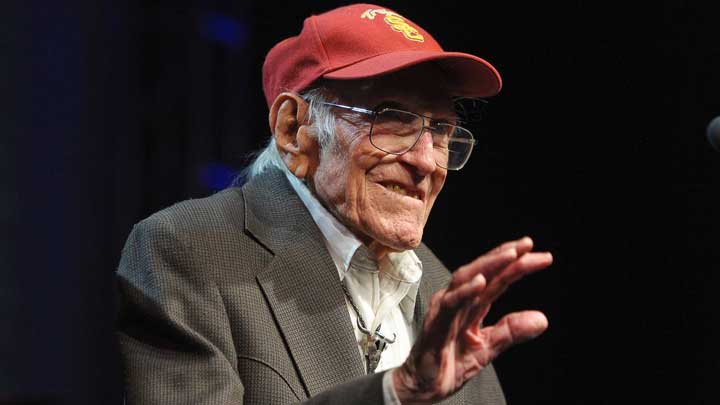 Louis Zamperini, wearing a red cap and suit, replying to a question on a public platform, symbolizing his resilience and triumph over challenges linked to Oppositional Defiant Disorder (ODD).