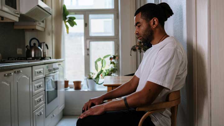 A man in his 30s with an unshaven face sitting on a chair, looking down with a sad and intense expression, symbolizing Major Depressive Disorder.