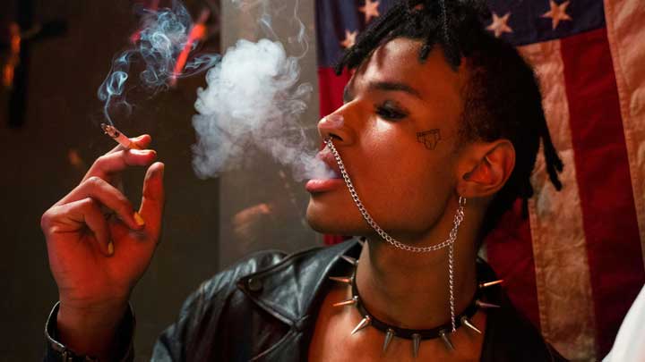 A young African-American man smoking, wearing a dog necklace, black leather jacket, and a silver chain from nose to ear, with a USA flag on the wall in the background.